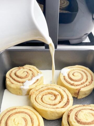 pitcher pouring cream over cinnamon roll dough in baking pan.
