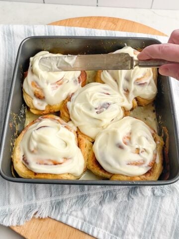 hand holding a knife spreading frosting on cinnamon rolls.