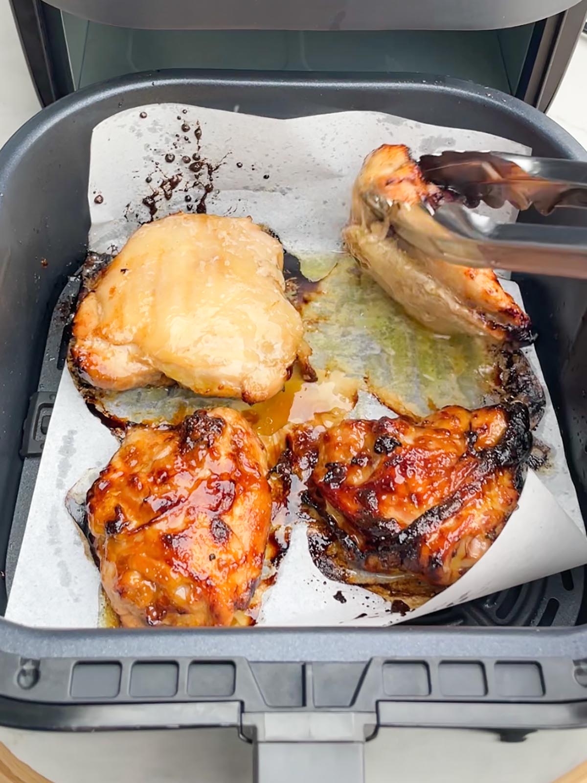 teriyaki chicken thighs in air fryer basket with tongs flipping one over