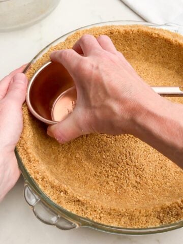 hand holding a measuring cup packing graham cracker crumbs into pie plate.
