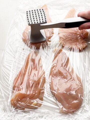 meat mallet pounding thin sliced chicken between layers of plastic wrap
