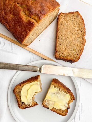 piece of banana bread cut in half spread with butter on a white plate.