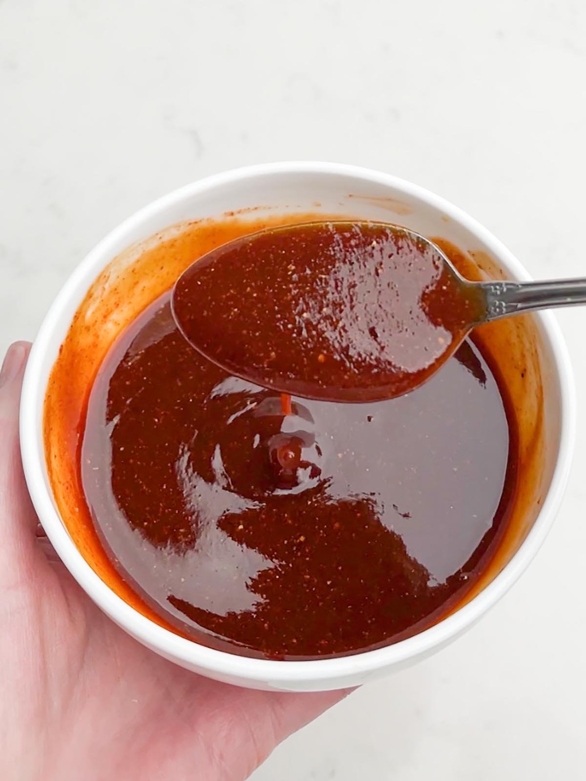 hand holding a white bowl of bbq sauce with a spoonful of sauce hovering over the bowl