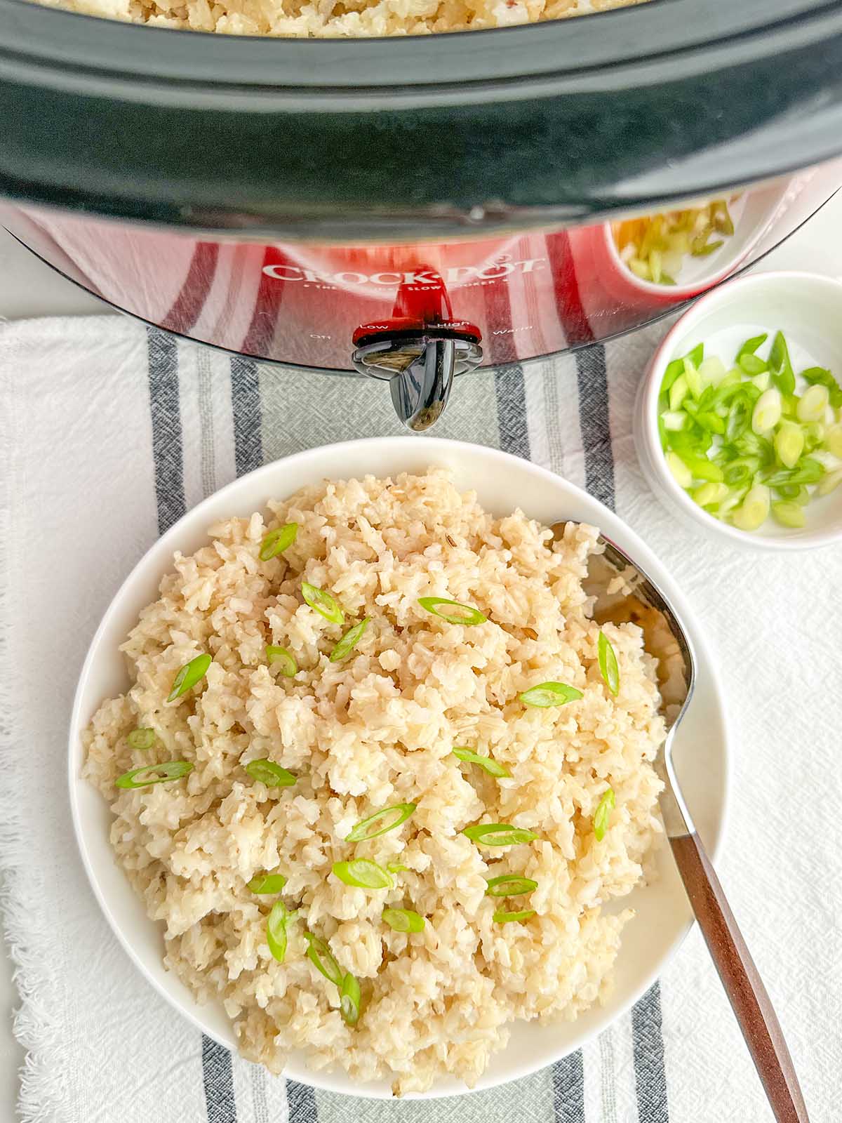 cooked brown rice and green onions in a white bowl by a red crock pot.