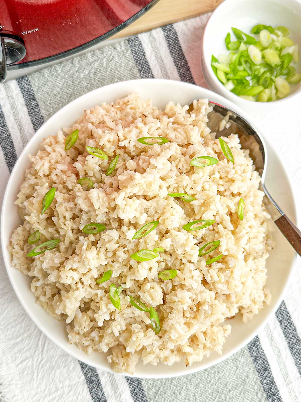 cooked brown rice and green onions in a white bowl by a red crock pot.