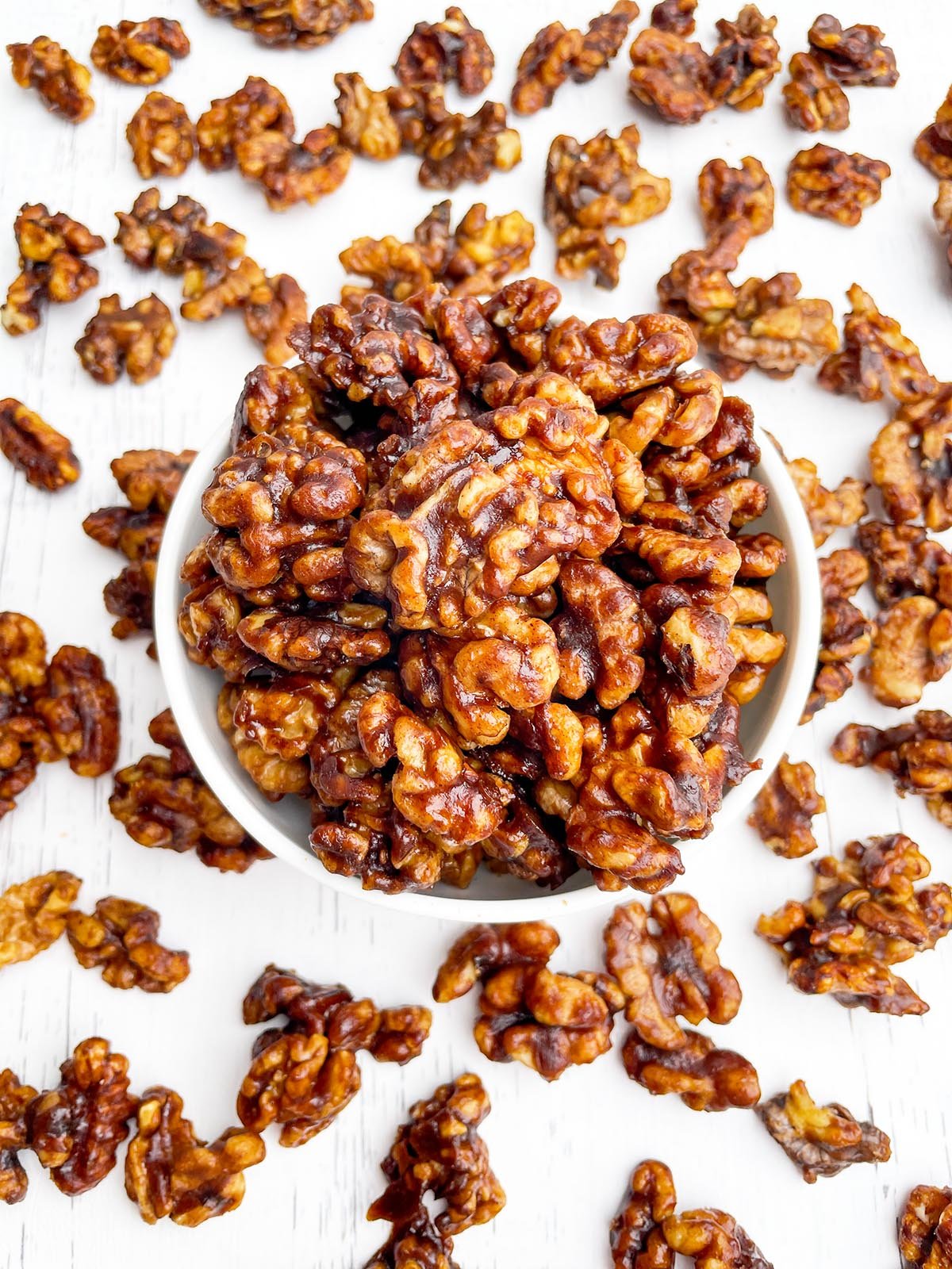 brown sugar candied walnuts in a white bowl on a surrounded by walnuts on a white countertop.