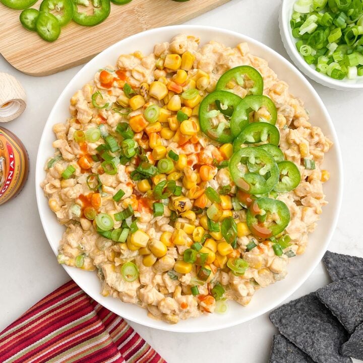 corn dip with cream cheese topped with corn, green onions, and jalapenos, and hot sauce in a white bowl