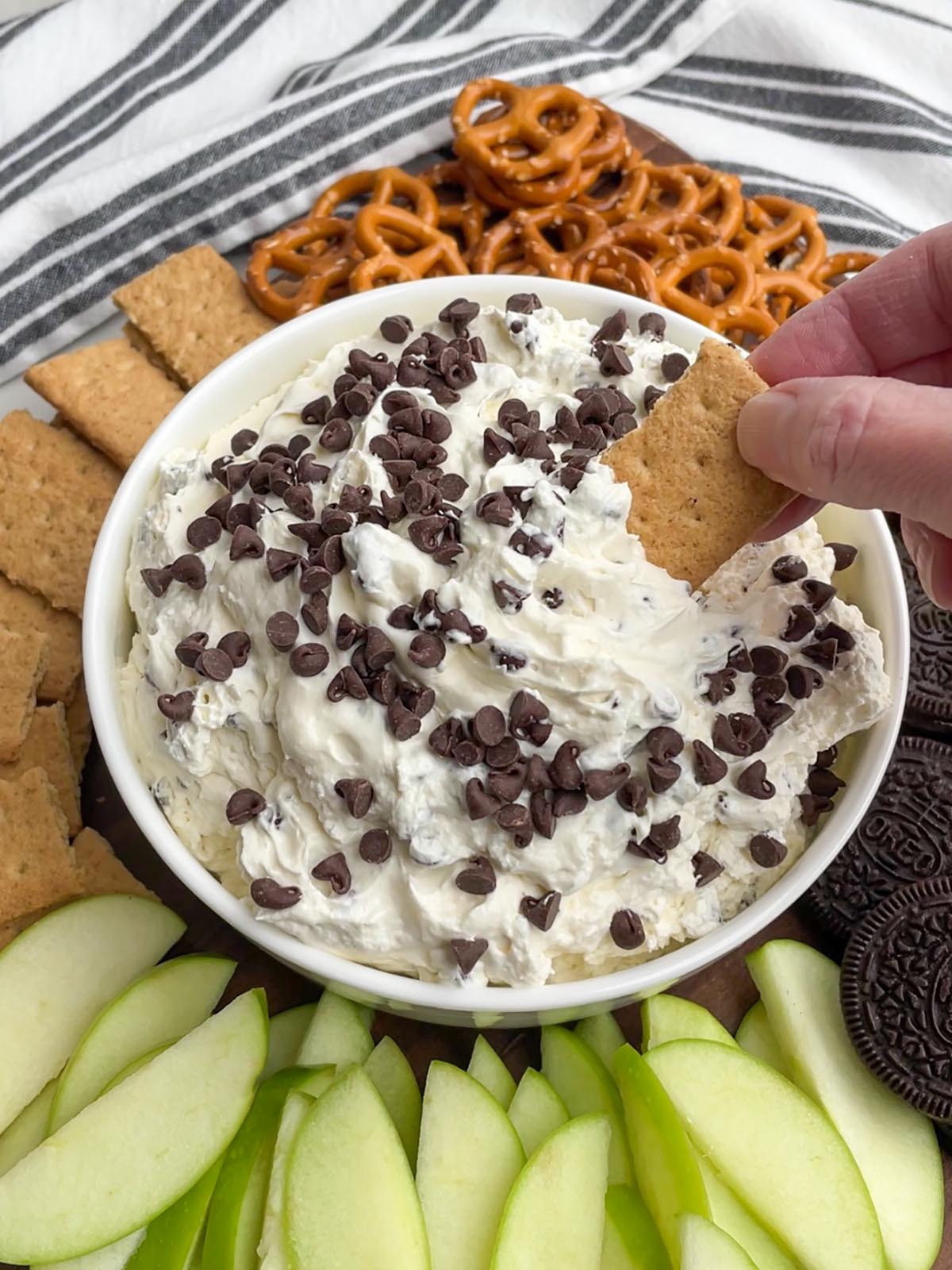 hand holding a graham cracker scooping into a white bowl of cream cheese chocolate chip dip with dippers in the background