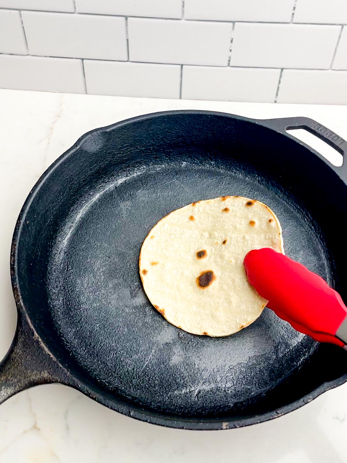 red tongs flipping a corn tortilla in a cast iron skillet