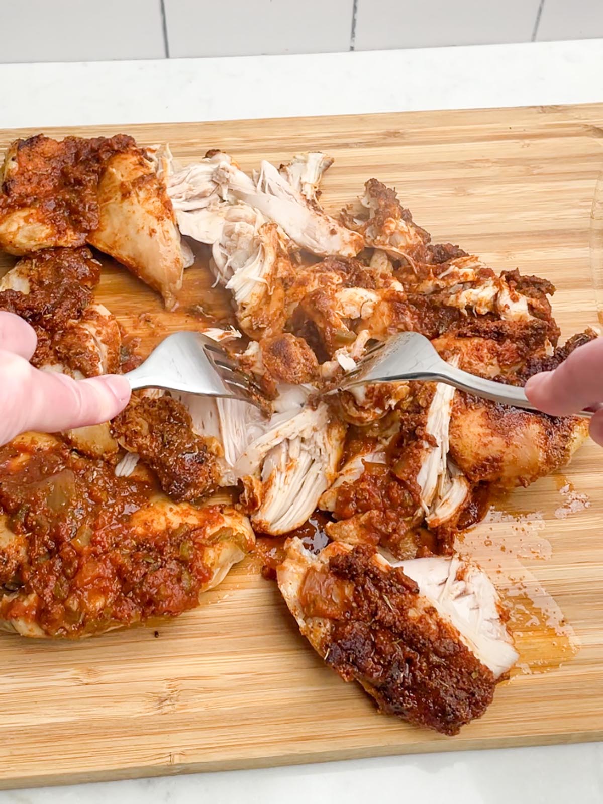 two hands holding forks shredding cooked salsa chicken on a wooden cutting board