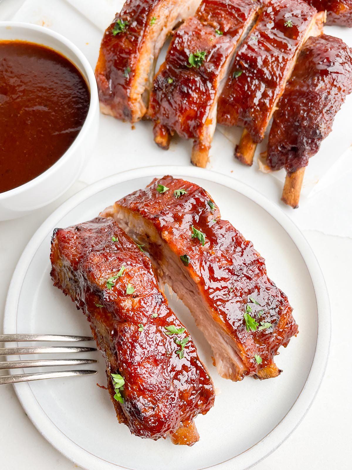white plate with two ribs with bowl of BBQ sauce and more ribs in the background.
