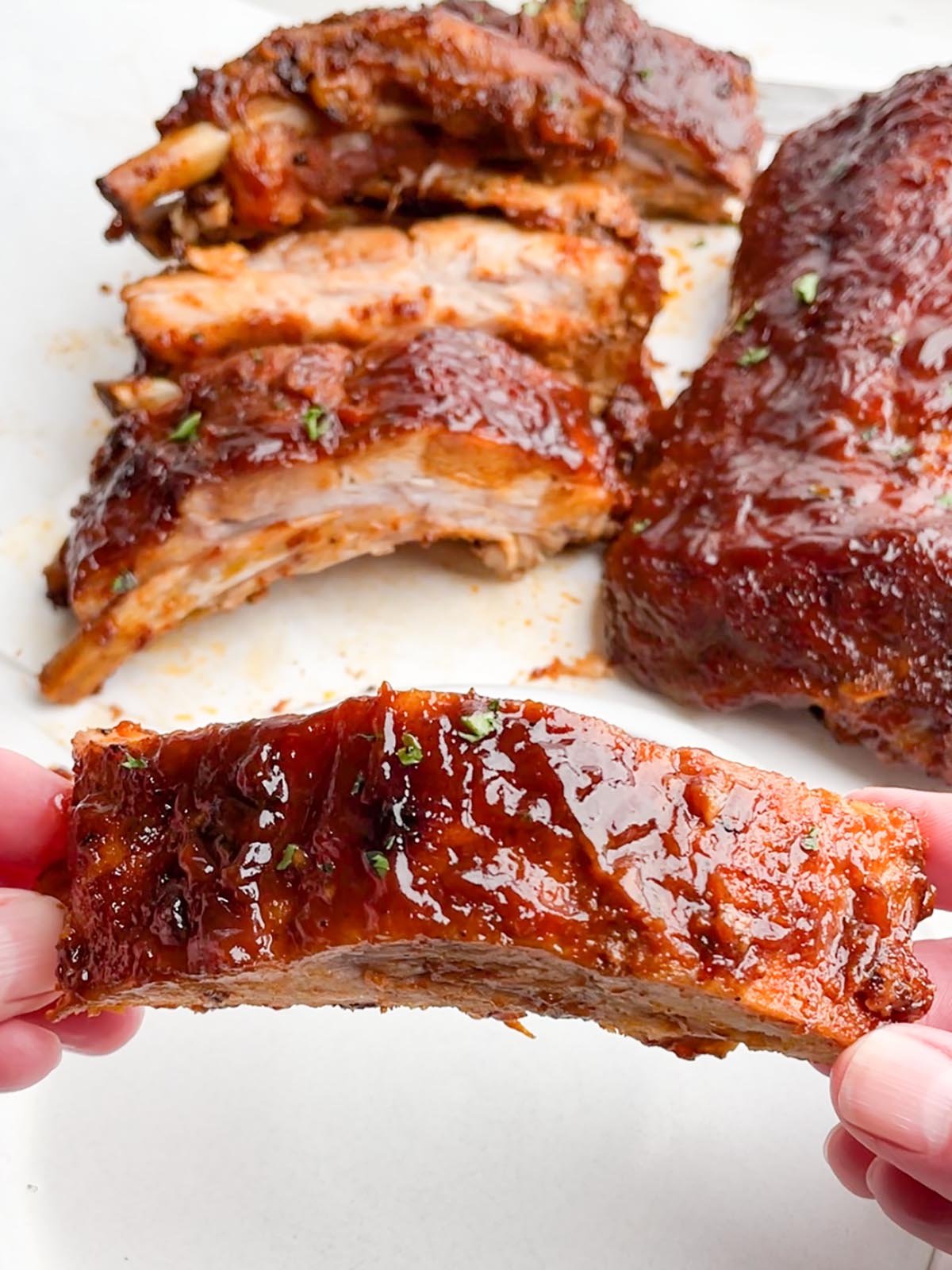 hands holding one rib in foreground with racks of ribs in the background.