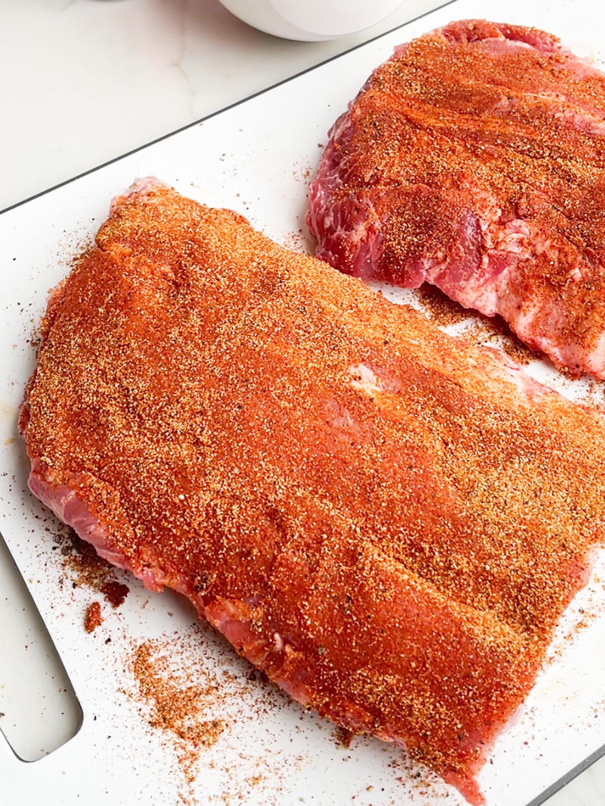 two half racks of ribs with dry rub on a white cutting board.