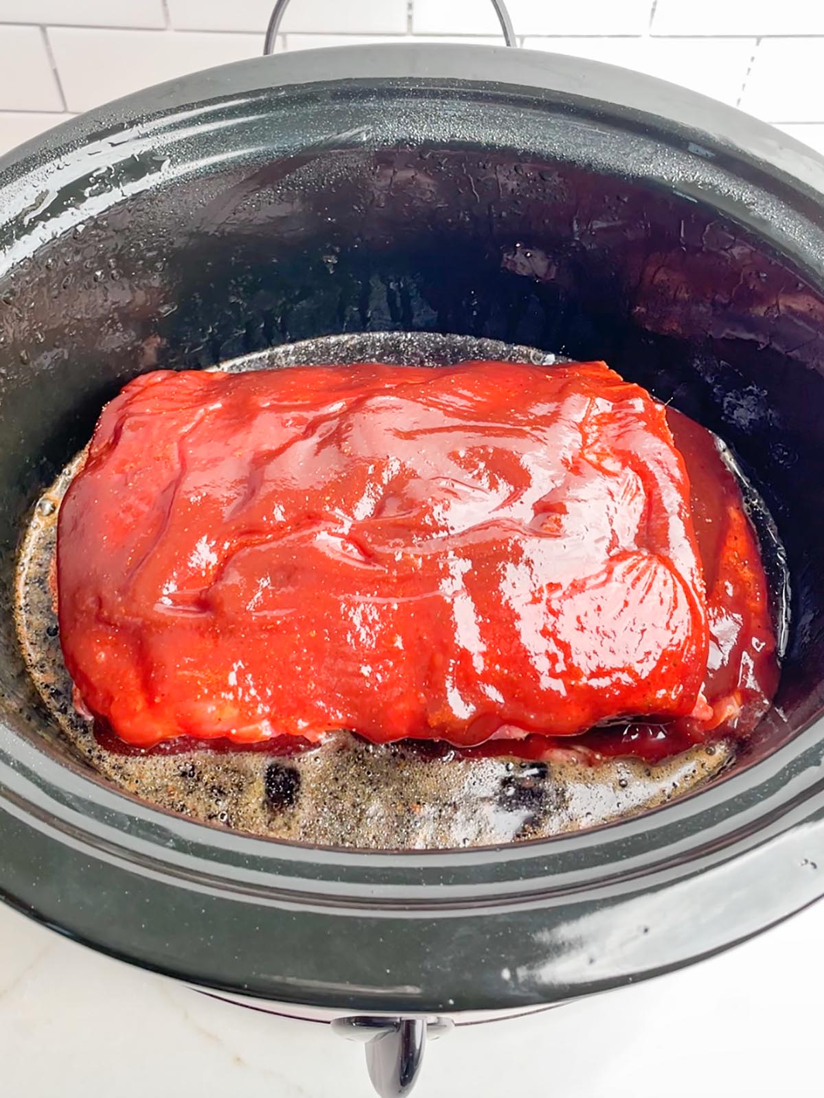 ribs with BBQ sauce in a black slow cooker with Dr. Pepper in the bottom.