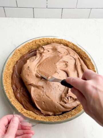 hand with spatula spreading creamy pudding layer over pie filling.