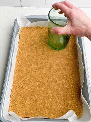 hand holding a glass pressing graham cracker crust in a 9 x 13 pan.