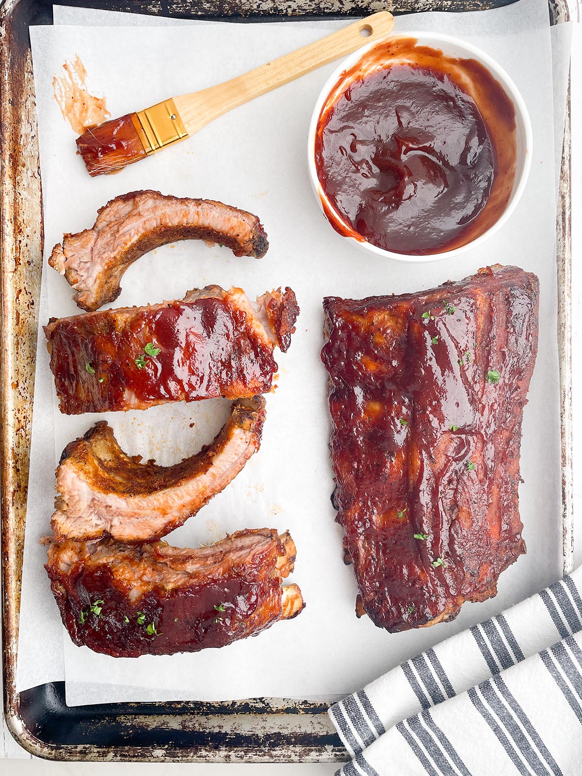one full rack of ribs and individual ribs on white parchment with a bowl of BBQ sauce and a paint brush
