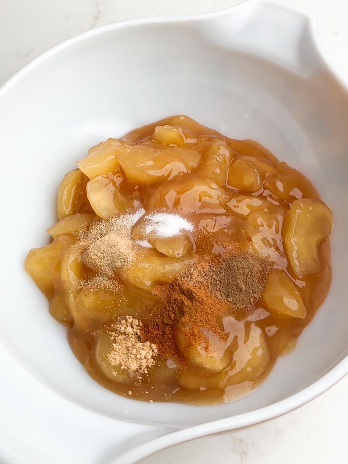Apple pie filling and spices in a white mixing bowl.