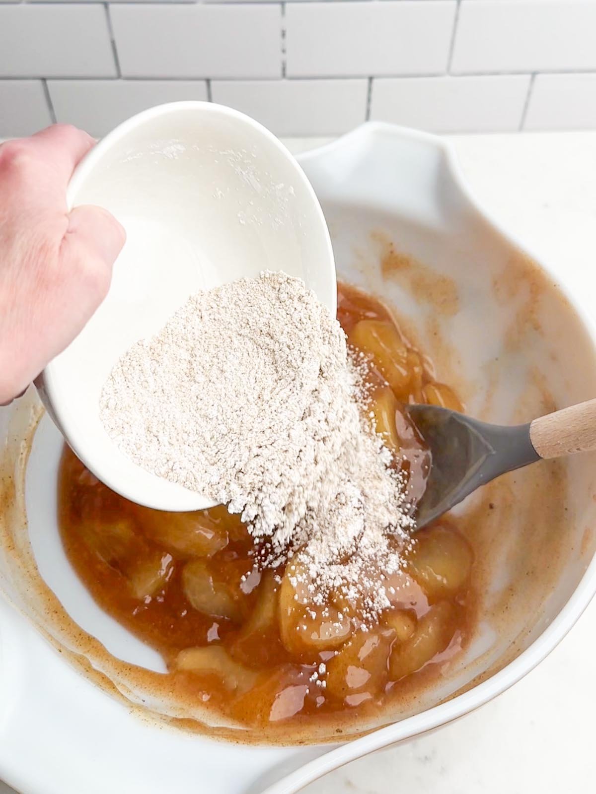 Hand pouring a bowl of cornstarch and brown sugar into apple pie filling.