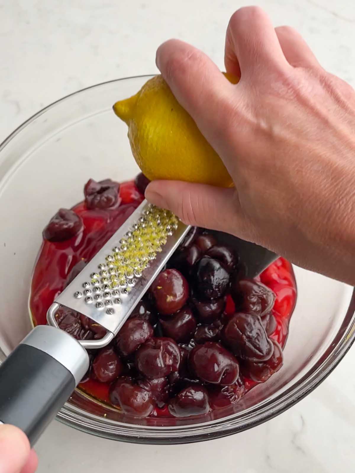 hand zesting a lemon with a microplane over a clear bowl of cherry pie filling