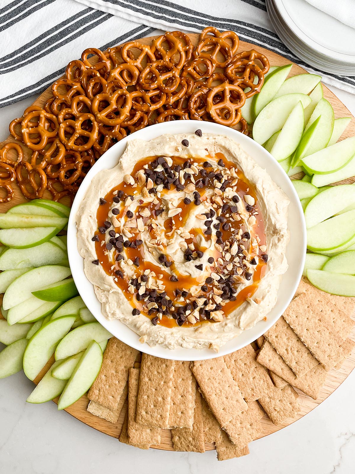 caramel apple dip in a white bowl on a round platter with pretzels, apples, and graham crackers