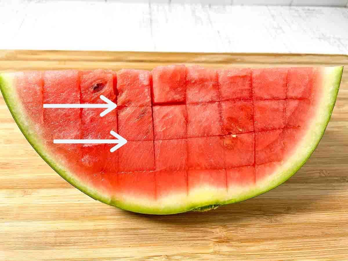 Watermelon quarter on a wooden cutting board with 2 white horizontal arrows