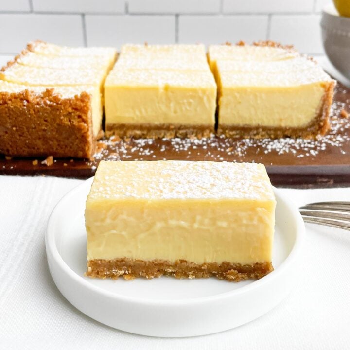 lemon bar with graham cracker crust on white plate with cutting board with bars in background