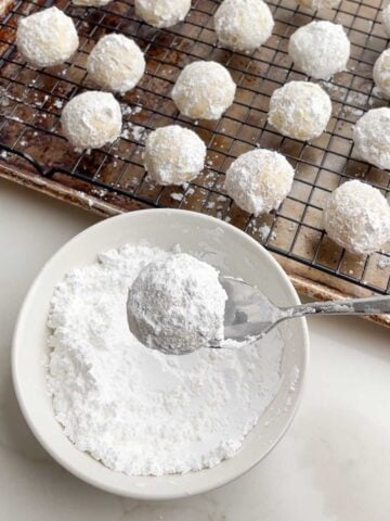 spoon lifting a rolled Mexican Christmas cookie out of a bowl of powdered sugar.