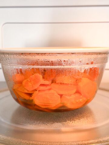 steamed carrots in a clear bowl covered with a plate in a microwave.