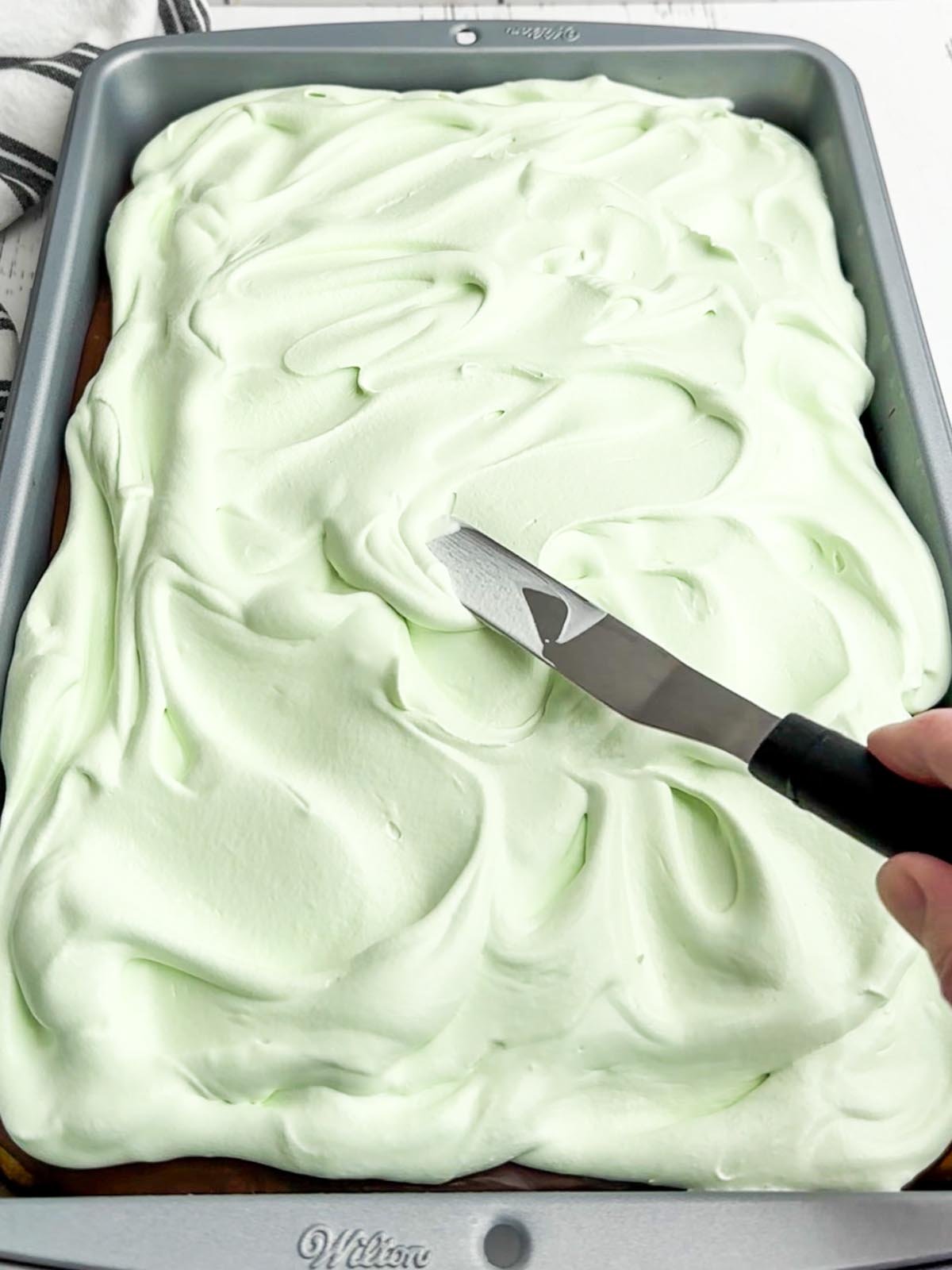 hand holding a spatula spreading green Cool Whip on top of cake