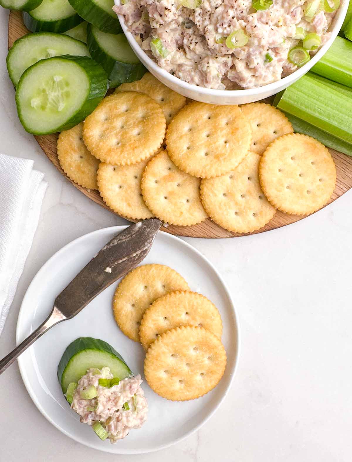 ham salad on a cucumber on a white plate with crackers next to a platter with ham salad, veggies, and crackers