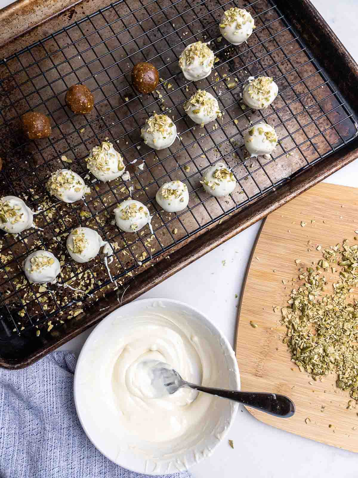pumpkin gingerbread truffles on a wire rack on a baking sheet next to a bowl of melted white chocolate and chopped pepitas