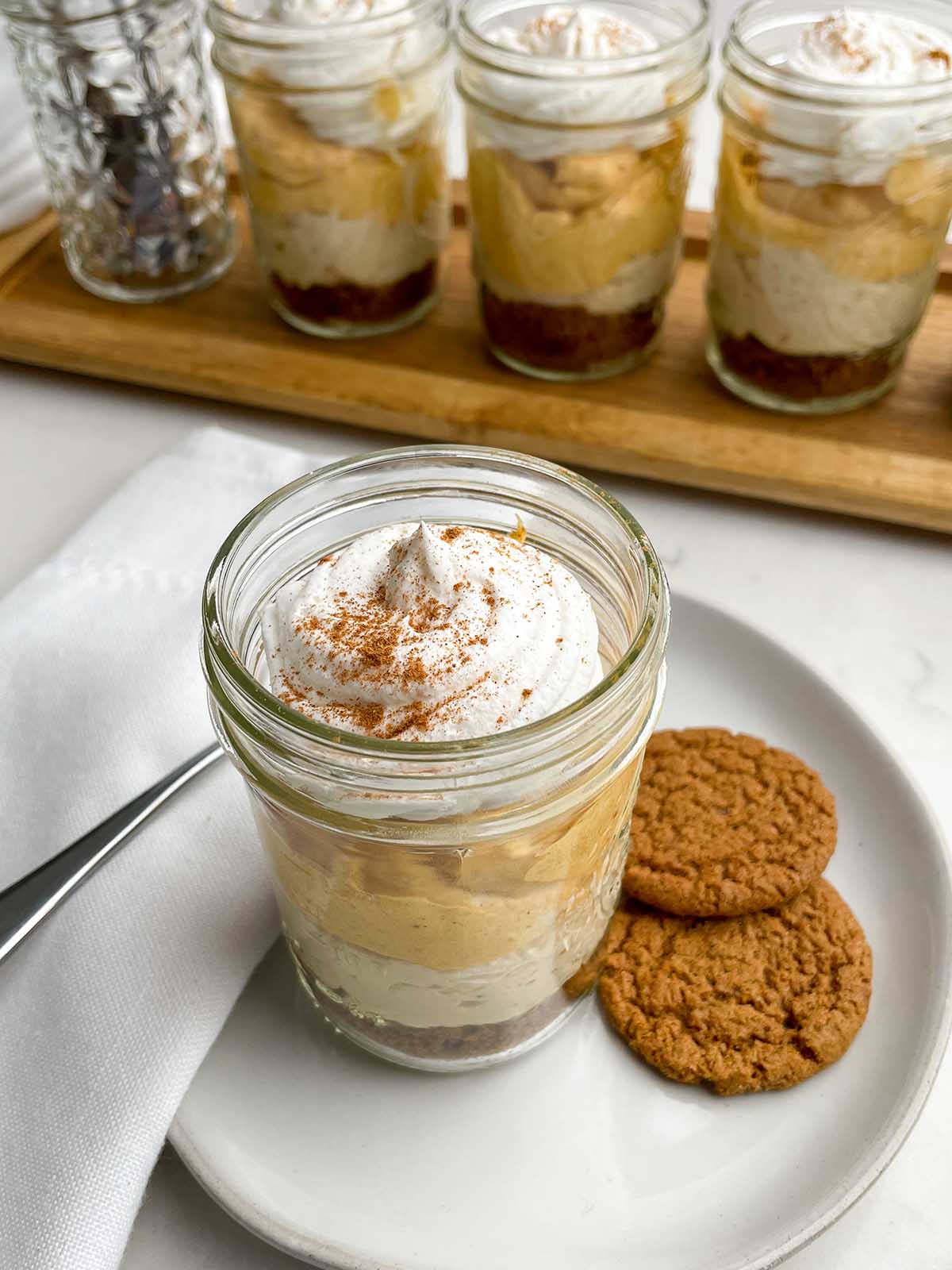a pumpkin pie in a cup on a plate with gingersnap cookies, a napkin, and a spoon in front of jars of pumpkin pie in a cup