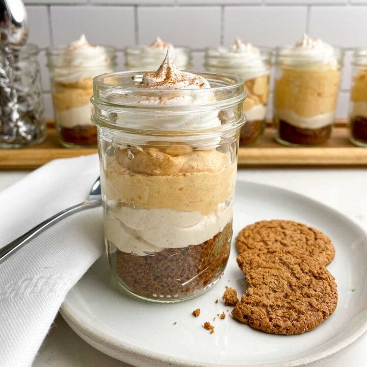 a pumpkin pie in a cup on a plate with gingersnap cookies, a napkin, and a spoon in front of jars of pumpkin pie in a cup