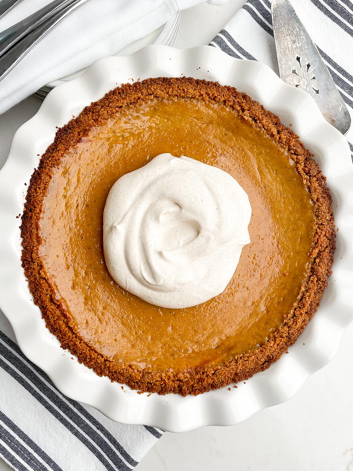 pumpkin pie with graham cracker crust in white pie plate with a dollop of cinnamon whipped cream in the center.