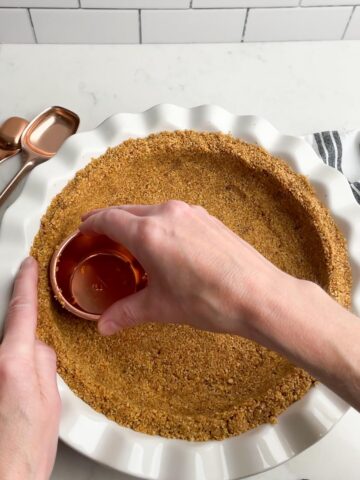 hand with measuring cup pressing graham cracker crumbs into pie plate.