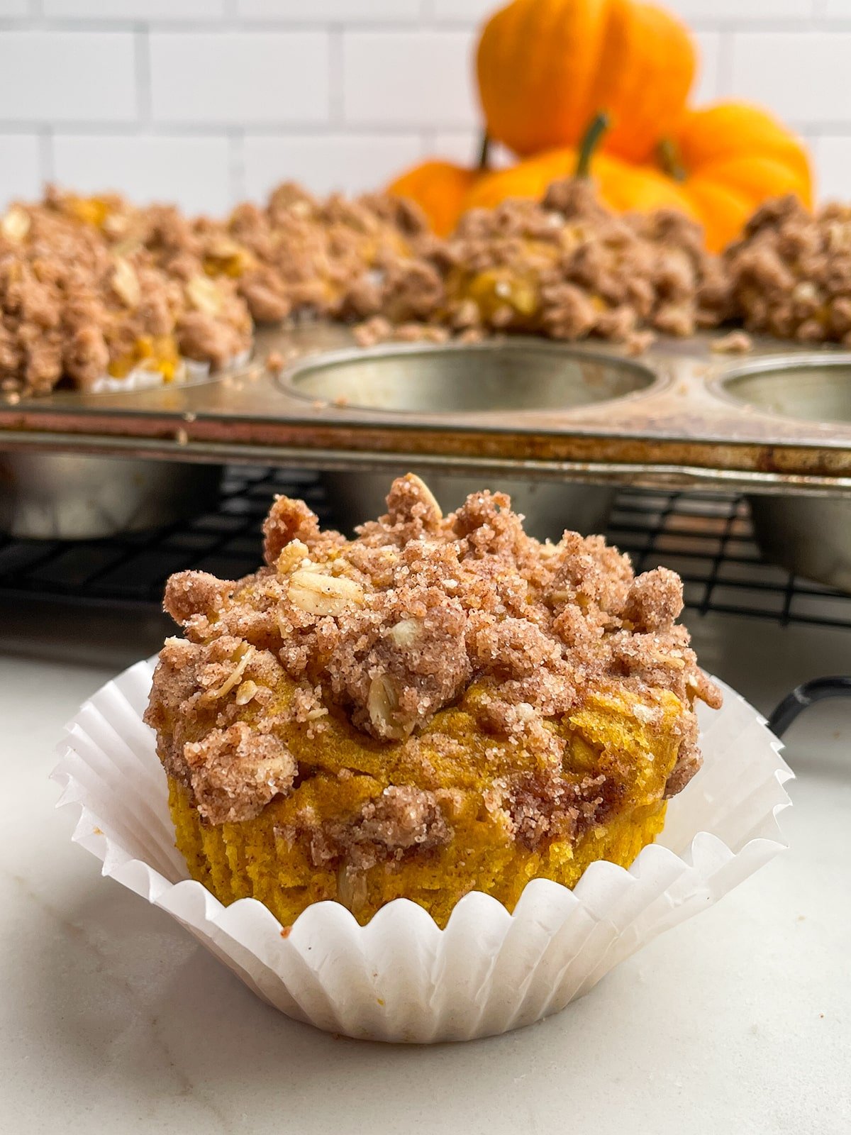 Pumpkin Snickerdoodle Muffin in a white paper muffin liner with pan of muffins in the background