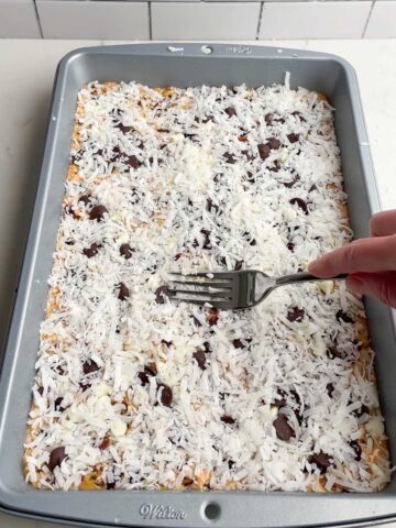 pumpkin magic bars in 9 x 13 pan with hand holding fork pressing down coconut.