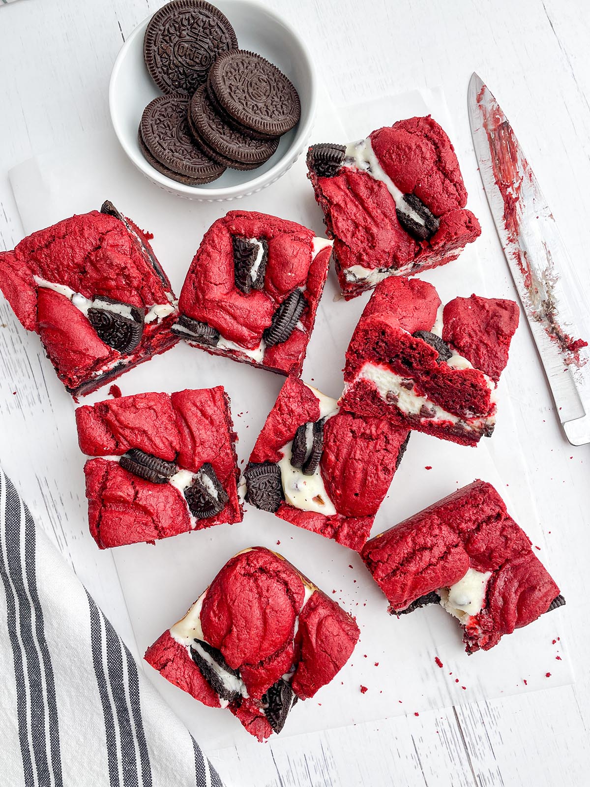 red velvet brownie squares on parchment paper next to a bowl of Oreos, a knife and a black and white napkin.