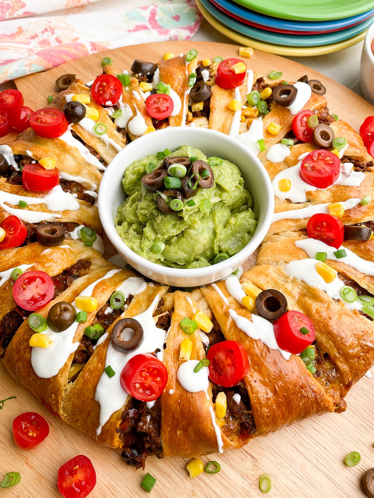 taco ring covered in toppings with guacamole in the center on a wooden cutting board