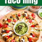 taco ring covered in toppings with guacamole in the center on a wooden cutting board