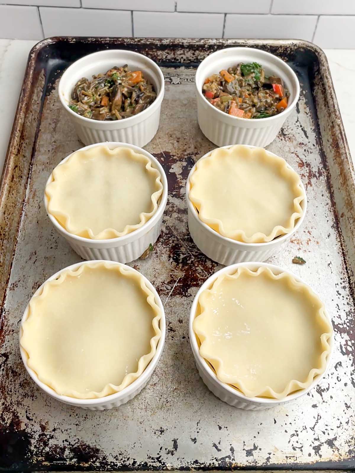 six ramkeins on a baking sheet filled with vegetarian pot pie filling with pie crust on four of them